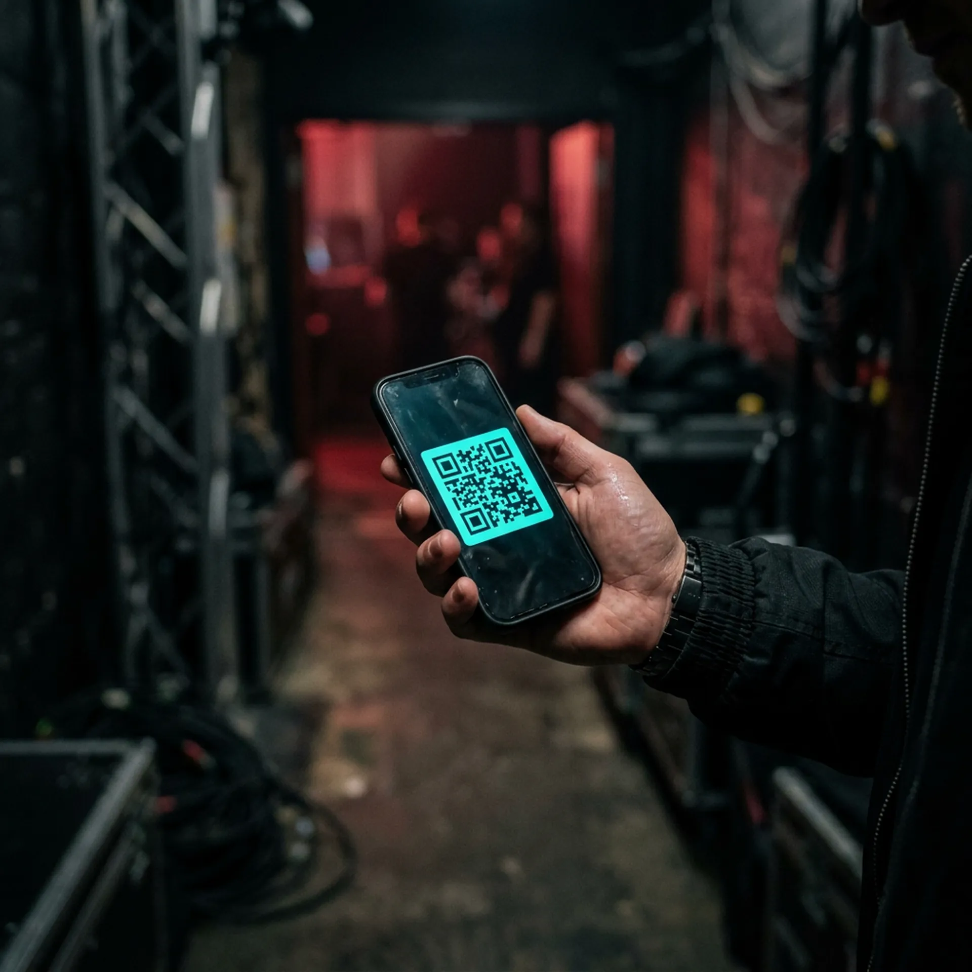 DJ hand holding smartphone displaying QR code in dark backstage corridor with subtle crimson stage light