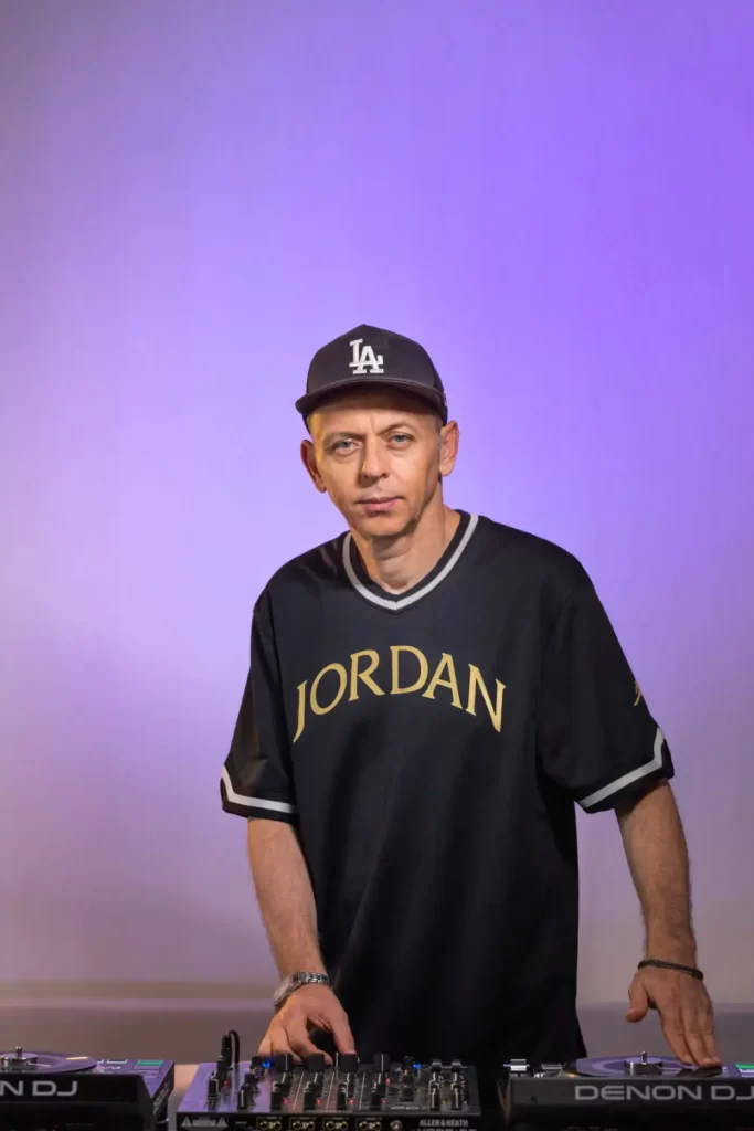 A man wearing a black "Jordan" shirt and a black cap stands behind DJ equipment against a purple-lit background, spinning the beats of DJ Efunk on The Fusion Radio.