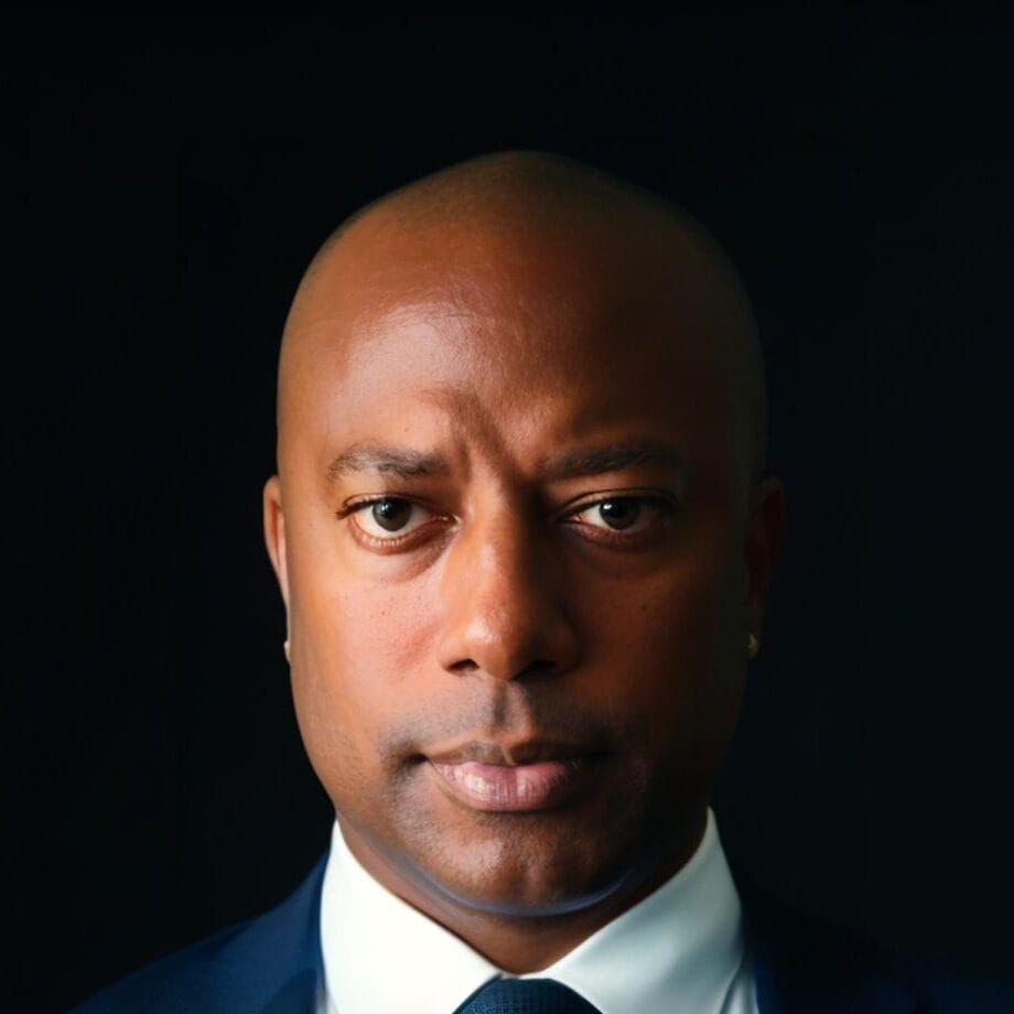 Scott Evans, a bald man in a dark blue suit, white dress shirt, and dark tie, is staring directly at the camera with a serious expression. The background is dark, emphasizing his formal attire and focused demeanor.