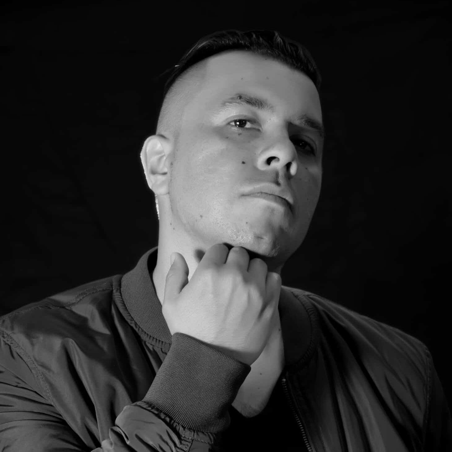 Black and white photo of DJ Puga, exuding confidence as they gaze at the camera. With short hair and a thoughtful hand on their chin, they wear a jacket against a dark backdrop, embodying the spirit of The Fusion Radio.